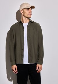 Olivově zelená košile na knoflíky, lehký materiál, černé knoflíky, nošena přes bílé tričko, doplněna béžovou kšiltovkou a černými kalhotami.