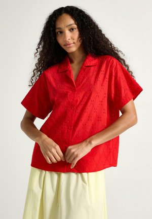Kvinde med langt krøllet hår iført en kortærmet rød øjetsbluse og lysegul nederdel, justerer blusens kant.