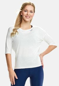 Bijela atletska majica s tri četvrtine rukava, mekani materijal, okrugli izrez i mali crni logotip na prsima, u kombinaciji s plavim tajicama.