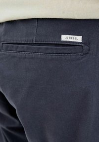 Κοντινό πλάνο πίσω τσέπης σκούρου ναυτικού μπλε παντελονιού με ραμμένη την ετικέτα της μάρκας JJ Rebel πάνω από αυτήν, φορεμένο με ανοιχτό μπεζ τοπ.