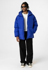Pufferjack in felblauw, met een capuchon, ritssluiting en elastische manchetten. Draag het over een witte shirt, gecombineerd met zwarte broeken en sneakers.