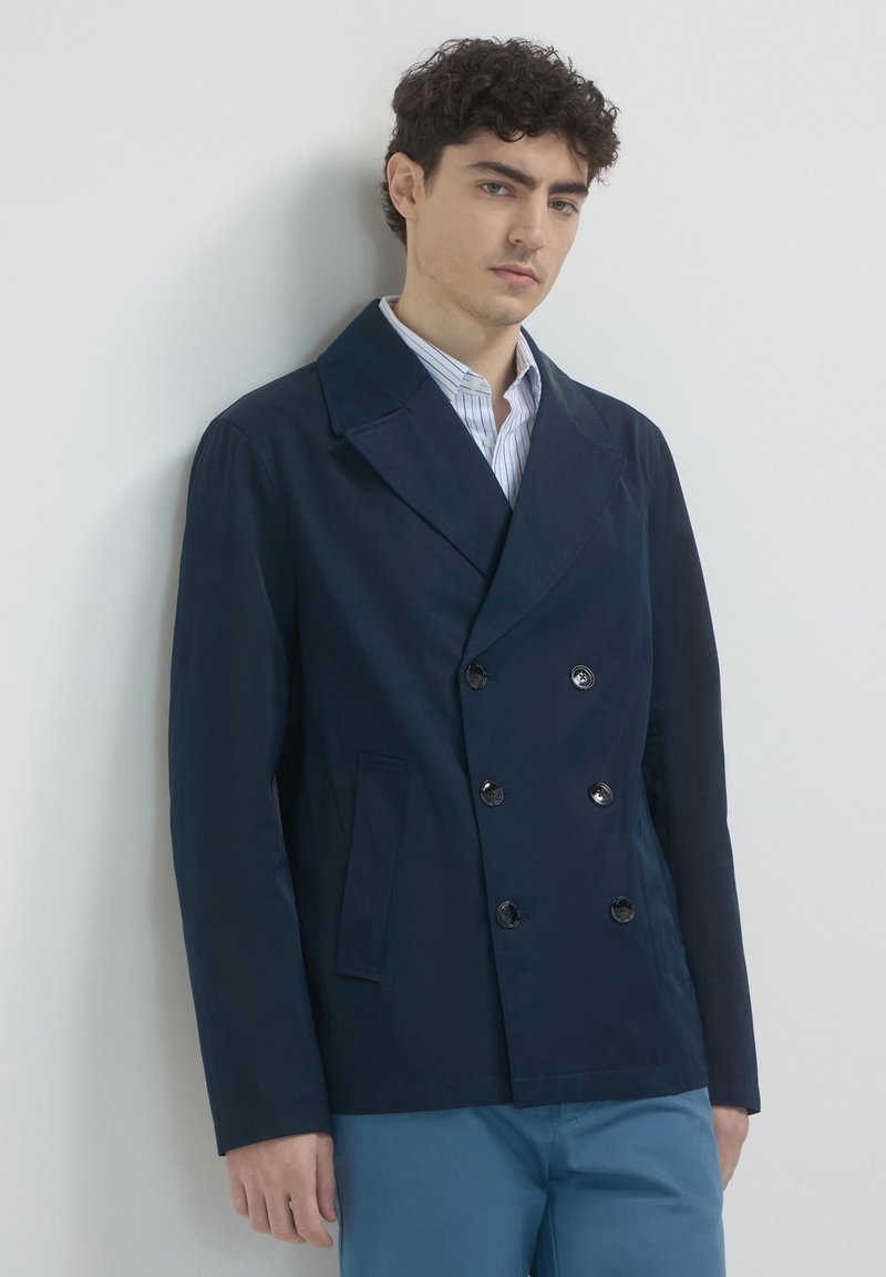 Giovane uomo con capelli ricci e scuri, vestito con un blazer blu navy doppiopetto, una camicia a righe e pantaloni blu, in piedi contro un muro bianco semplice.