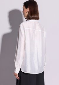 Biała koszula z długim rękawem wykonana z lekkiej tkaniny o gładkiej fakturze, z zaokrąglonym dołem i ozdobnymi mankietami. Posiada szew na plecach w formie yoke.