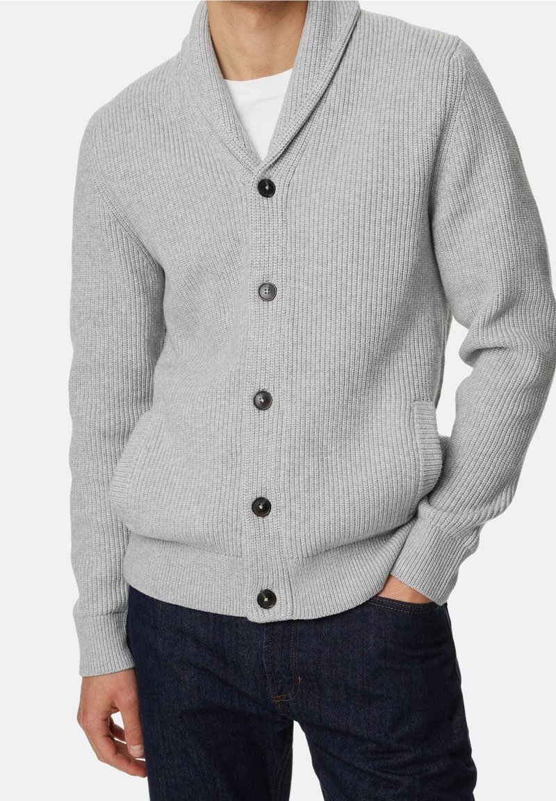 Homme portant un cardigan en maille côtelée gris clair avec des boutons noirs sur une chemise blanche et un jean bleu foncé, une main dans la poche.