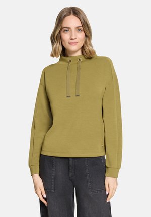 Frau trägt ein olivgrünes Sweatshirt mit hohem Kragen und Kordelzug sowie schwarze Jeans und steht vor einem schlichten hellen Hintergrund.