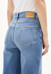 Lichtblauwe denim jeans met een middelhoge taille. Vierkante achterzakken en een bruine leren patch met het opschrift "ARMEDANGELS" in gouden letters.