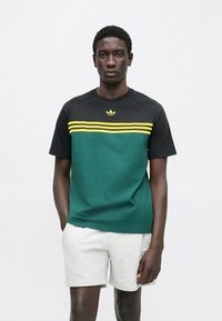 Žalios ir juodos spalvų trumpomis rankovėmis marškinėliai su geltonomis juostelėmis ir logotipu ant krūtinės, derinami su šviesiai pilkomis kelnėmis. Lygios audinio tekstūros.