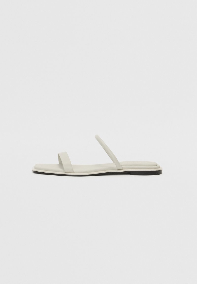 Sandale minimaliste blanc cassé avec deux fines brides et une semelle plate sur un fond uni gris clair.