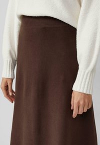 Bruine gebreide rok met een zachte textuur, met een hoog uitgesneden tailleontwerp, gecombineerd met een witte geribde trui. Close-up van handen op de rok.