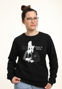 Vrouw met een bril en haar in een knot draagt een zwarte trui met een gesplitst zwart-wit Addams personage dat een boek en een zwaard vasthoudt.