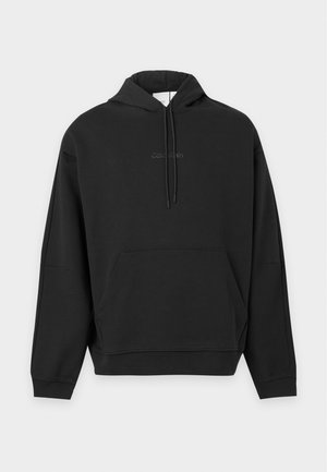 Ung mand iført en sort Calvin Klein hættetrøje med frontlomme og snore, kombineret med mørkegrå jeans, stående mod en ensartet baggrund.