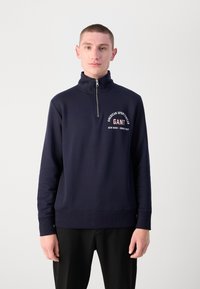 Navy blue zip-up sweatshirt with a high collar, featuring a white graphic print that reads "GANT," and ribbed cuffs and hem.