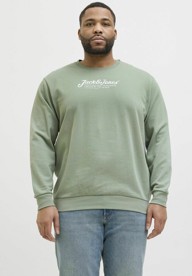 Grüne Sweatshirt aus weichem Stoff, mit Rundhalsausschnitt und langen Ärmeln. Weißer Logotext auf der Brust, kombiniert mit hellblauen Jeans.