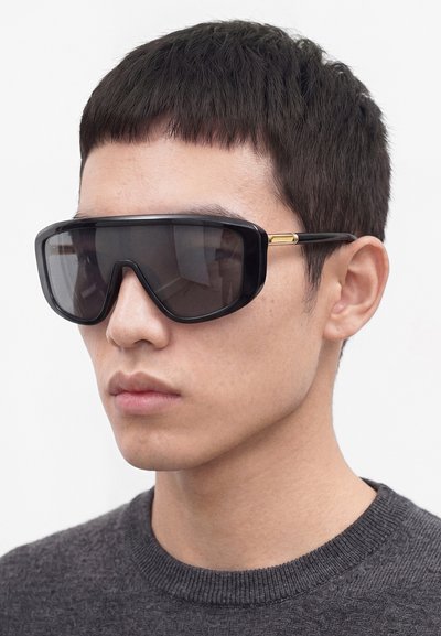 Jeune homme aux cheveux courts et foncés portant de grandes lunettes de soleil enveloppantes noires et un pull gris foncé à col rond sur un fond uni.