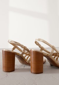 Gold metallic heeled sandals with a strappy design. Features a wooden block heel and angled straps, showcasing a smooth texture.