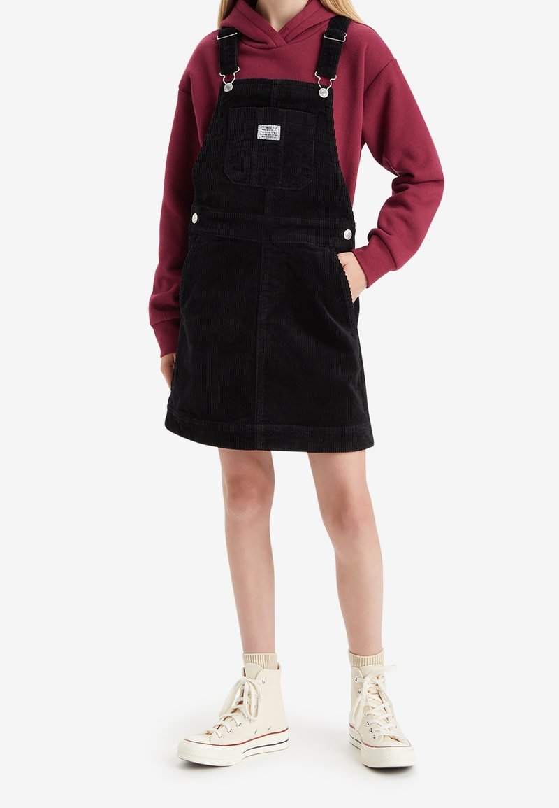 Personne portant une robe salopette en velours côtelé noire par-dessus un sweat à capuche bordeaux et des baskets montantes blanches avec des chaussettes.