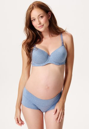 Gravid kvinde med langt brunt hår iført lyseblå amme-bh og matchende undertøj foran en ensfarvet baggrund.