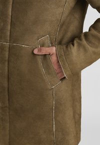 Brauner Wildledermantel mit einer Vordertasche, aus der eine sichtbare Hand herausragt, kontrastierenden Nähten und einer strukturierten Oberfläche. Elegantes, minimalistisches Design.