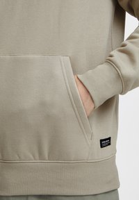 Beige sweatshirt met een voorzak, ribgebreide manchetten en tailleband. Heeft een klein zwart label nabij de zaknaad. Zachte textuur.