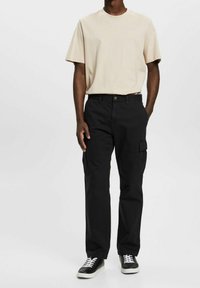 Beige t-shirt med rund hals, svarta cargo-byxor med sidofickor och svarta sneakers med vita snören. Outfiten har en avslappnad passform.