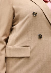 Μπεζ σακάκι κουστούμι με δύο μεγάλα μαρμάρινα κουμπιά και τσέπη με καπάκι, φορεμένο πάνω από ροζ ένδυμα.