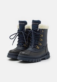 Bottes d'hiver bleu marine avec col en fausse fourrure, œillets en métal et système de laçage. Semelle en caoutchouc avec texture antidérapante et détail de logo sur le côté.