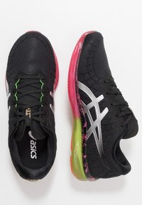 Zapatillas deportivas negras con parte superior texturizada, detalles plateados y una suela multicolor en rosa y verde. Presentan cordones neón en contraste.