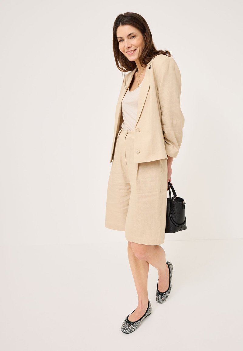 Conjunto de blazer de lino beige y pantalones cortos, combinado con una camiseta sin mangas clara. Un bolso negro y unas bailarinas negras con estampado completan el atuendo.