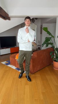 Νέος άνδρας με λευκό πουλόβερ ζιβάγκο και χακί παντελόνι στέκεται σε σαλόνι με πορτοκαλί καναπέ, μεγάλο φυτό, ξύλινο πάτωμα και τηλεόραση στο φόντο.