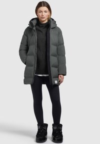 Parka imbottita grigio scuro con cappuccio, dotata di chiusura con zip e tasche laterali, indossata sopra un maglione nero e leggings neri, abbinata a stivali neri.