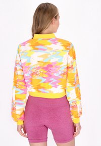 Geknipte jas met een helder, abstract patroon in geel, roze en blauw. Heeft een gele kraag en rekbare manchetten. Gecombineerd met roze shorts.