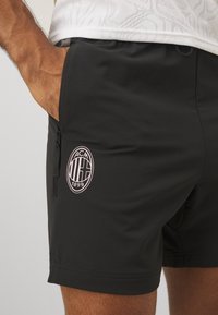 Shorts de sport noirs avec un logo ACM rose sur le côté gauche, et une poche zippée à droite. Tissu lisse, design ajusté.