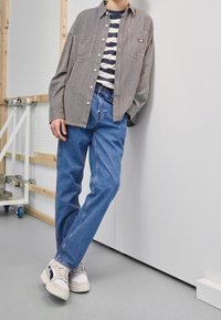 Camicia a righe bianca e nera sopra una t-shirt a righe blu e bianche, abbinata a jeans azzurri chiari e sneakers bianche, appoggiata a un muro.