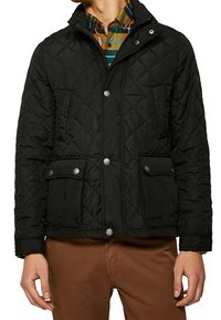 Giacca nera trapuntata con bottoni a pressione, tasche laterali e colletto, indossata sopra una camicia a quadri. Il materiale sembra liscio e isolante.