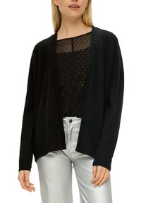 Schwarze Strickjacke mit offenem Vorderteil, geripptem Muster und langen Ärmeln, kombiniert mit einer transparenten schwarzen Bluse und metallisch silbernen Hosen.