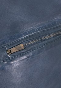 Blaues Ledertaschenabschnitt mit einem Reißverschluss und metallischem Ziehstück, glatter Textur und subtilen Falten, die auf ein weiches, angenehmes Material hinweisen.