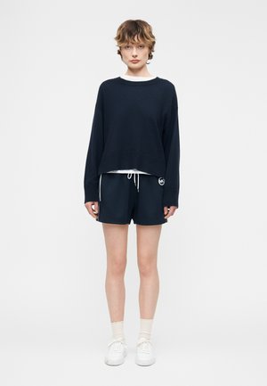 Mladá osoba stojaca, oblečená v čiernom nadrozmernom svetri, čiernych šortkách so šnúrkou, bielych ponožkách a bielych teniskách, neutrálnym pozadím.