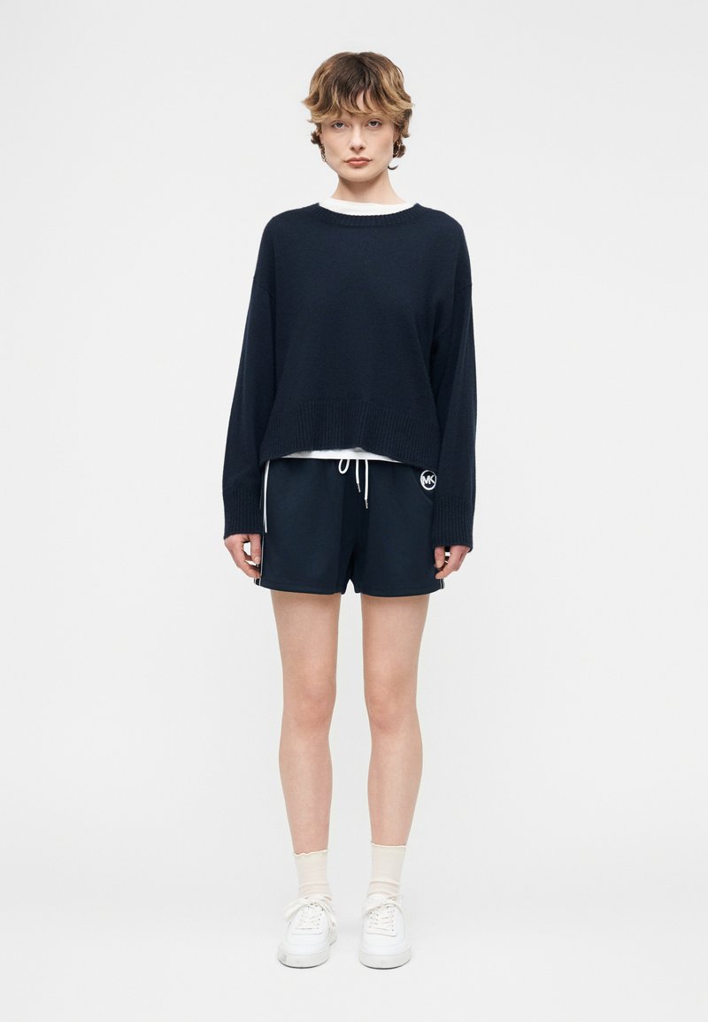 Νεαρό άτομο που στέκεται, φοράει μαύρο φαρδύ πουλόβερ, μαύρο σορτς με κορδόνι, λευκές κάλτσες και λευκά αθλητικά παπούτσια, ουδέτερο φόντο.