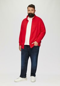 Rode zip-up jas over een witte shirt, gecombineerd met donkere spijkerbroek en witte sneakers. De jas heeft een hoge kraag en een losse pasvorm.
