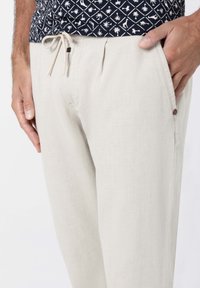 Helle beige Hosen aus strukturiertem Stoff, mit einem Kordelzugbund und seitlichen Taschen. Ein kleines Logo-Detail ist in der Nähe der Tasche sichtbar.
