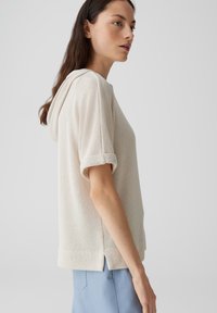 Pullover leggero beige con cappuccio, maniche corte arrotolate e spacchi laterali. Il tessuto testurizzato aggiunge dettaglio; design casual adatto per il layering.