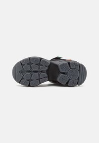 Suola in gomma nera con design testurizzato, caratterizzata da un diverso modello di grip e un logo al centro. Sono visibili accentuazioni di colore vivaci.