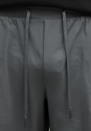 Graue Sportshorts werden angezeigt, mit einem Bund mit Kordelzug und schwarzen Spitzen. Sie sind von der Taille abwärts vor einem neutralen Hintergrund zu sehen.