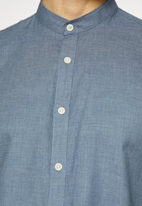 Camicia in chambray blu con colletto alla mandarino, bottoni bianchi, tessuto strutturato e sottile motivo intrecciato. Inquadratura ravvicinata della patta anteriore.