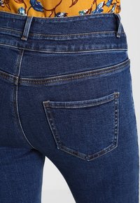 Vaqueros de un azul oscuro con tiro alto, diseño de cinco bolsillos, detalles de costura sutiles y una textura de denim suave.