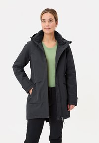 Waterdichte zwarte parka met een ritssluiting aan de voorkant, trekkoord capuchon, twee voorzakken en een zachte binnenvoering, gedragen met een groene top.