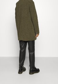 Persona vistiendo un abrigo largo texturizado verde oscuro, pantalones de cuero negros y botas de cuero negro con cordones, de pie frente a un fondo blanco liso.