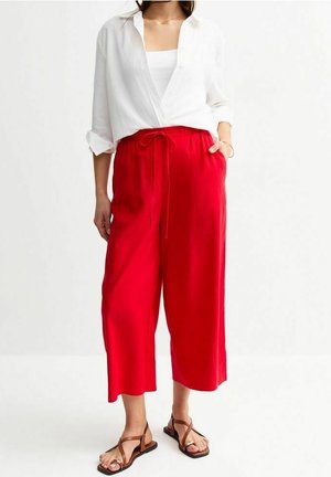 Vrouw draagt felrode, wijd uitlopende cropped broek, wit overhemd over een witte top en bruine sandaal met bandjes, staand tegen een witte achtergrond.