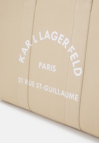 Kreemi kangas kott, millel on valge trükitud tekst: "KARL LAGERFELD PARIS 21 RUE ST-GUILLAUME." Tekst on ümberringi ja paksude tähtedega.