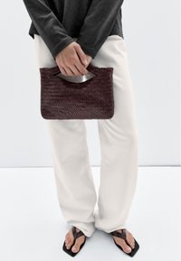 Borsa clutch intrecciata bordeaux con manico superiore, forma rettangolare, finitura testurizzata, abbinata a pantaloni bianchi e ciabatte nere.
